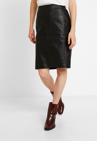 Jupe crayon en cuir noir avec une texture lisse, longueur genou, présentant des coutures verticales et une taille élastique. Portée avec des bottines bordeaux brillantes.