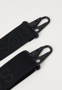 Sangle en tissu noir avec deux clips en métal. La texture est tissée, avec des lettres en relief sur un côté. Les clips ont une finition mate.