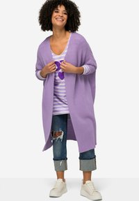 Ljuslila stickad cardigan med öppen fram, bärs över en randig lila skjorta med ett hjärtmotiv, i kombination med slitna jeans och vita sneakers.