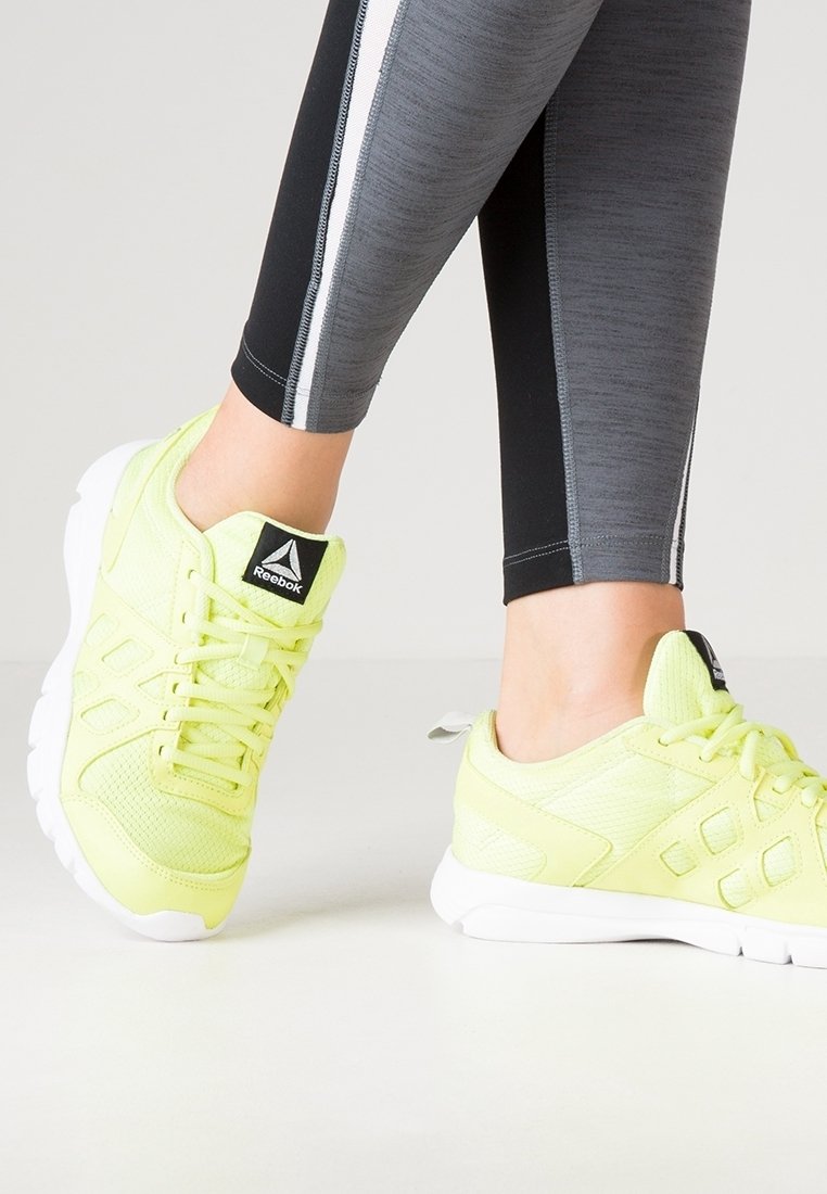 Neon-gelbe Sportschuhe mit Mesh-Obermaterial, weißer Sohle und schwarzem Reebok-Logo. Kombiniert mit schwarzen und grauen Leggings mit weißen Streifen.