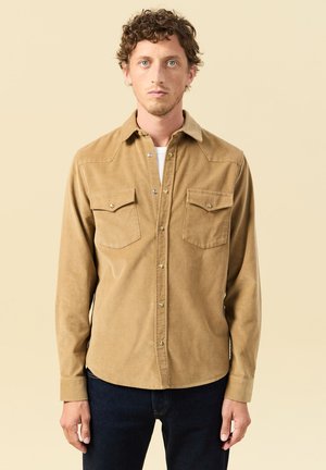 Junger Mann mit lockigem Haar, der ein beige Knopfleistenhemd aus Cord über einem weißen Oberteil und dunklen blauen Jeans trägt, vor einem beigen Hintergrund stehend.