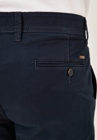 Marineblaue Hose aus einem glatten, gewebten Stoff mit einer Gesäßtasche mit Knopfverschluss und einem kontrastierenden Stich entlang des Taillenbunds.