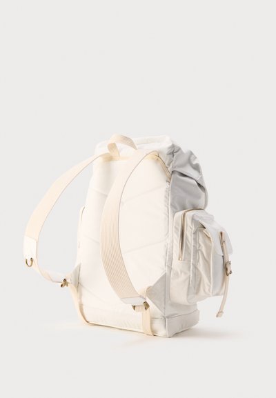 Κρεμ και ανοιχτό γκρι σακίδιο με επενδυμένους ιμάντες και μια μπροστινή τσέπη με φερμουάρ, φαίνεται από πίσω σε απλό φόντο.