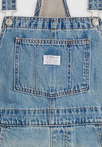 Τζιν φορμάκι σε ανοιχτό μπλε με ρυθμιζόμενους ιμάντες. Διαθέτει τσέπη στο στήθος με ετικέτα και εμφανείς ραφές. Ομαλή υφή.