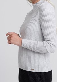 Jasnoszary sweter z prążkowanej tkaniny, z wysokim kołnierzem i długimi rękawami. Ma subtelne detale w haftach oraz mały złoty element dekoracyjny.