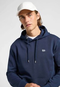 Granatowa bluza z kapturem z kieszenią kangurzą, regulowanym sznurkiem i srebrnymi końcówkami. W połączeniu z białą czapką z subtelnym logo.