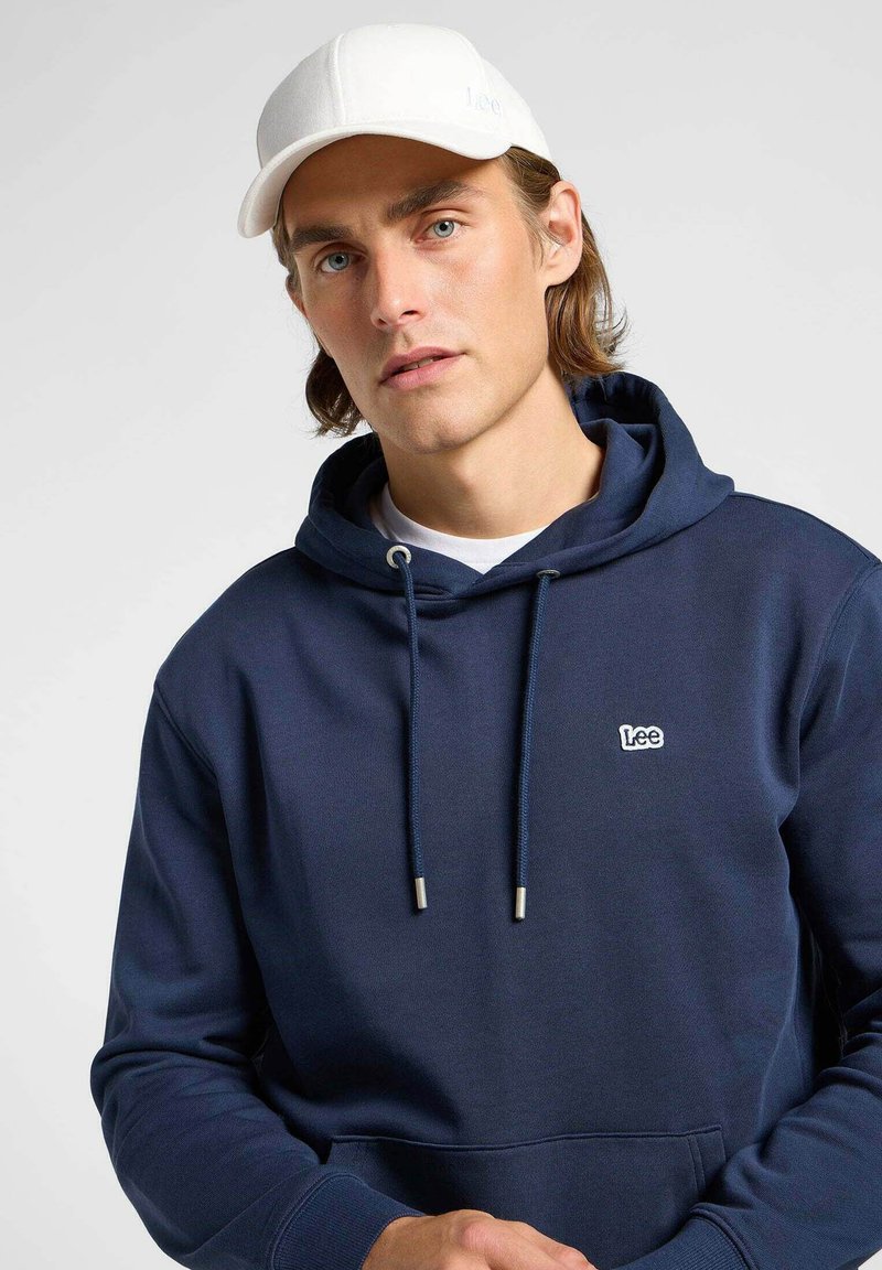 Granatowa bluza z kapturem z kieszenią kangurzą, regulowanym sznurkiem i srebrnymi końcówkami. W połączeniu z białą czapką z subtelnym logo.