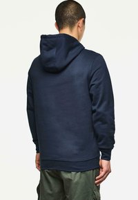 Uomo che indossa un felpa blu navy e pantaloni cargo verdi, in piedi con le spalle alla camera su uno sfondo bianco uniforme.