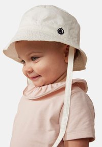 Cappello a secchiello bianco con una leggera lucentezza, presenta un emblema di una barca a vela blu navy e lacci bianchi. Indossato con una camicia rosa chiaro con colletto arricciato.