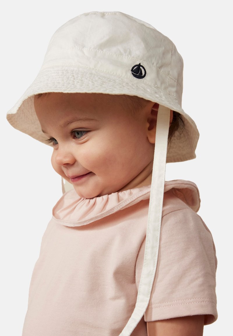 Cappello a secchiello bianco con una leggera lucentezza, presenta un emblema di una barca a vela blu navy e lacci bianchi. Indossato con una camicia rosa chiaro con colletto arricciato.