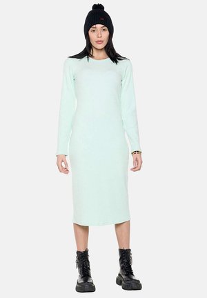 Femme portant une robe midi bleu clair à manches longues, un bonnet en maille noir, des bottes noires épaisses à lacets et des bracelets dorés, debout devant un fond blanc.