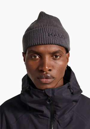Mand med mellemmørk hud iført en grå ribstrikket hue med "HALO" broderi og en sort lynlåsjakke med høj krave.