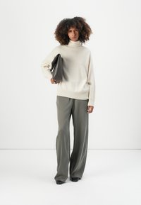 Helles beige Rollkragensweatshirt mit geripptem Saum, kombiniert mit lockeren, olivgrünen Hosen. Das Model hält eine dunkelgraue, strukturierte Schultertasche.