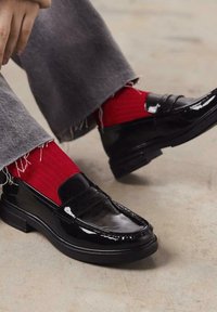 Mocassins en cuir verni noir au design épuré, avec un bout arrondi et un détail de sangle discret, associés à des chaussettes rouges côtelées.