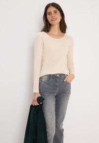 Haut beige à manches longues associé à un jean gris ; le mannequin tient une veste vert foncé. Tissu doux avec un col rond et une coupe ajustée.