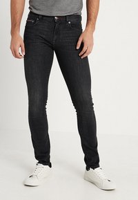 Svarta slim fit-jeans med en slät yta, metallknapp och röda sömmar, kombinerat med vita sneakers och en grå tröja.
