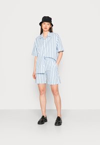 Camisa de manga curta azul e branca com riscas e calções a condizer, feitos de um tecido leve, combinados com um chapéu preto à bucket e sapatos pretos.