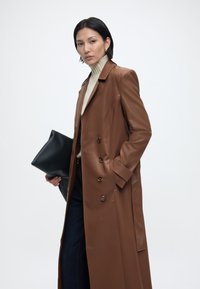 Γυναίκα με μακρύ καφέ δερμάτινο παλτό, εκρού ζιβάγκο, σκούρα τζιν, κρατώντας μεγάλο μαύρο φάκελο, στέκεται μπροστά σε λευκό φόντο.