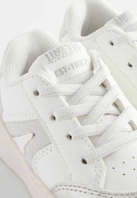 Baskets blanches en cuir et en mesh. Design à lacets, avec une semelle texturée et un détail de logo sur le côté "Établi en 1982."