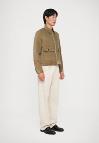 Brūns korduroja žakets ar rāvējslēdzēju priekšā un divām krūšu kabatām, apvienots ar krēmkrāsas platām biksēm un melniem mokasīniem.