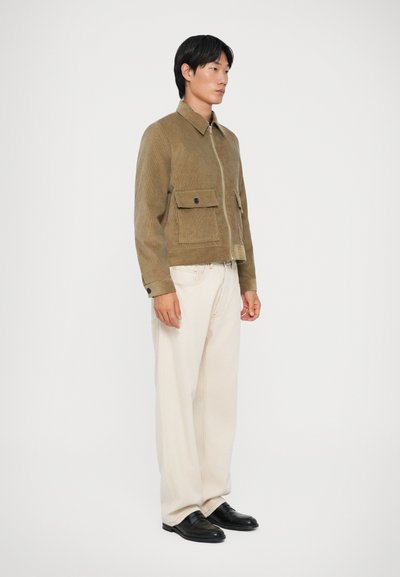 Brun fløjlskajak med lynlås foran og to brystlommer, parret med cremede vidde bukser og sorte loafers.