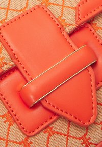 Pomarańczowy pasek ze skóry z złotą metalową klamrą, zamocowany na beżowej tkaninie z pomarańczowym wzorem geometrycznym.