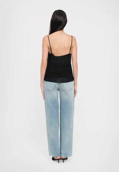 Camisole en dentelle noire avec dos en dentelle et fines bretelles, associé à un jean large taille haute bleu clair et des talons noirs, vu de derrière.