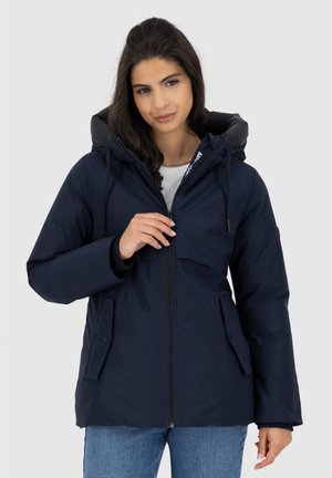 Giacca isolante blu navy con cappuccio, dotata di chiusura con zip, due tasche frontali e polsini elastici. Realizzata in un tessuto morbido e resistente alle intemperie.