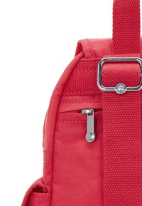 Dettaglio di uno zaino in tessuto rosso che presenta una tasca laterale con zip, una tracolla testurizzata e accessori in metallo con un motivo a stella sul tirante della zip.