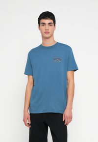 Blå bomulds-T-shirt med korte ærmer, rund halsudskæring og en lapdetalje med et grafisk design på venstre bryst.