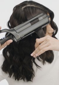 Femme en train de boucler ses cheveux foncés avec un fer à friser triple baril noir, tenant une mèche de cheveux d'une main et le fer à friser de l'autre.