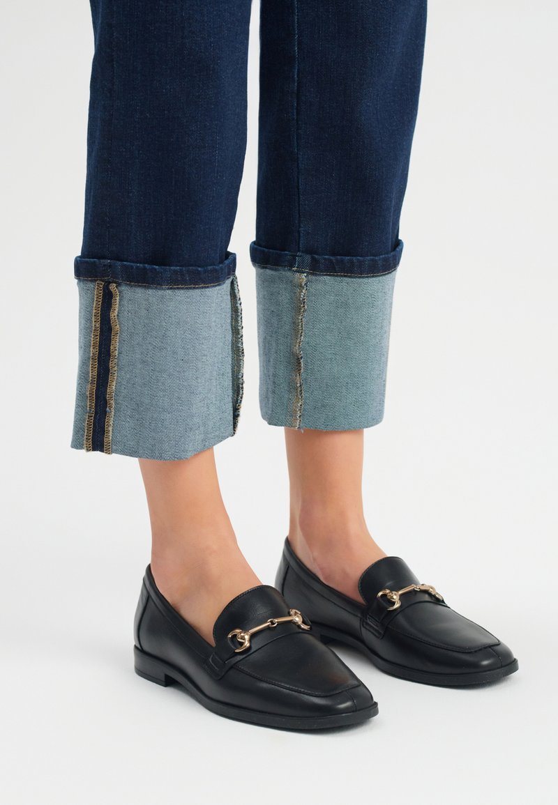 Zwarte leren loafers met gouden hardware-detail, gecombineerd met donkerblauwe denim jeans met een contrasterende lichtblauwe zoom en een geFranjeerd zoom.