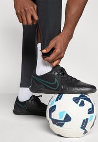 Sorte Nike fodboldsko med teal detaljer, tekstureret overdel og gummisål placeret over en mønstret hvid og blå fodbold.