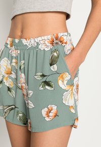 Shorts floraux en tissu vert avec des fleurs orange et blanches, taille élastique, poches latérales et une coupe décontractée avec un ourlet légèrement effiloché.