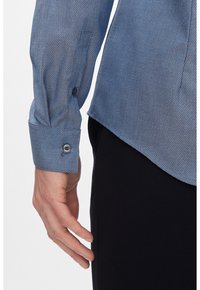 Camicia blu con motivo, realizzata in tessuto testurizzato, con polsini a bottoni e orlo curvo. Una mano è visibile mentre tiene il bordo della camicia.