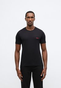 Černé tričko s krátkým rukávem z bavlny. Má kulatý výstřih a malý červený nápis „HUGO“ na levé straně hrudi.