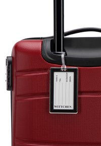Red hard-shell suitcase with textured surface, retractable handle, and a tag holder displaying a name and contact information label.