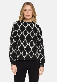 Vrouw in een zwart lange mouw trui met een wit geometrisch diamantpatroon en zwarte broek, staande tegen een effen achtergrond.