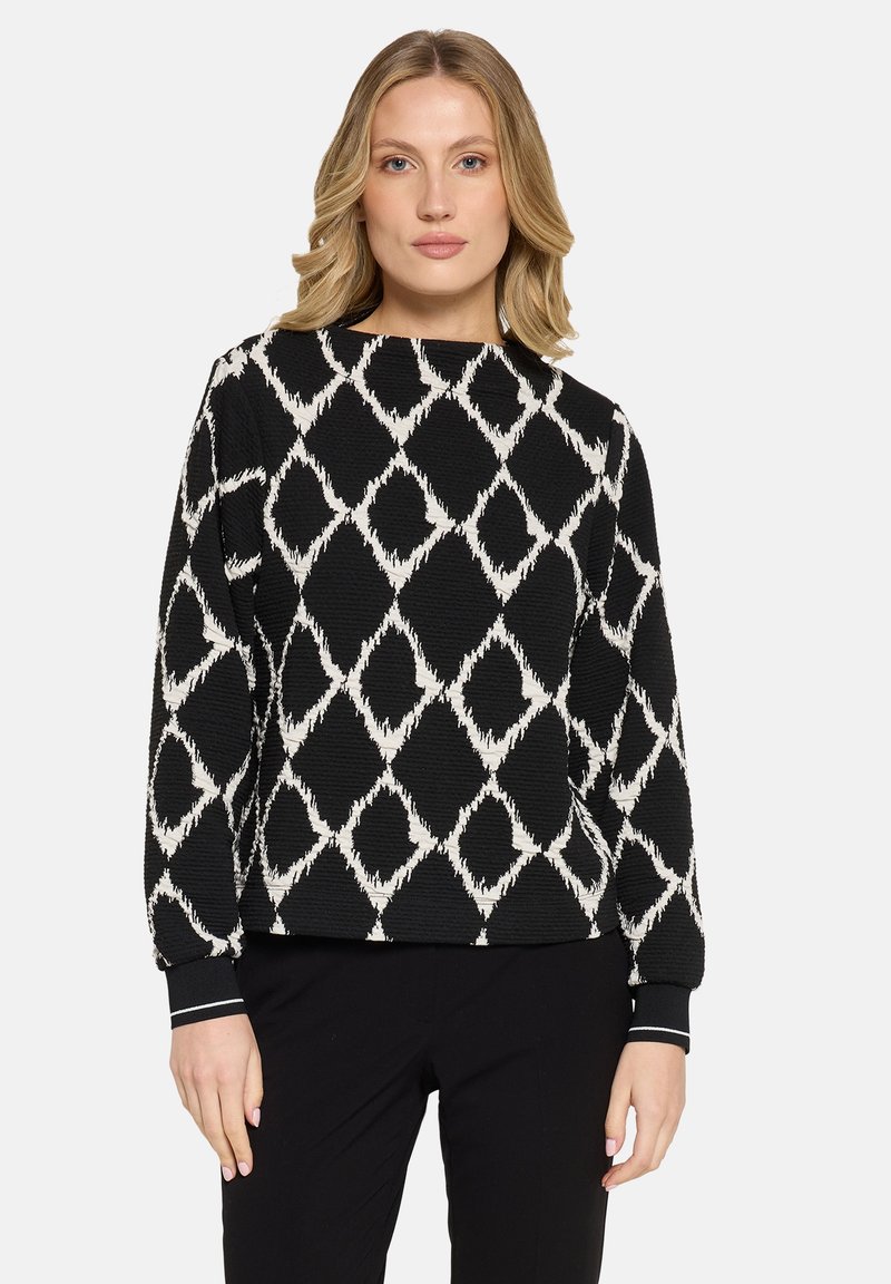 Vrouw in een zwart lange mouw trui met een wit geometrisch diamantpatroon en zwarte broek, staande tegen een effen achtergrond.