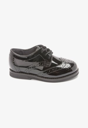 Chaussure brogue en cuir verni noir avec fermeture à lacets et perforations décoratives, vue de côté sur fond blanc.