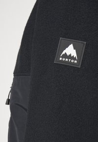 Czarna kurtka z polaru o teksturowanej powierzchni, czarnym panelu materiałowym oraz kwadratowej naszywce z logo przedstawiającym górę i napisem "BURTON".