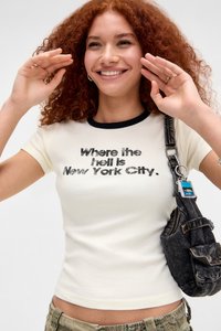 Krämfärgad t-shirt med korta ärmar och svart ribbat halsband, med fet text som lyder "Var i helvete är New York City." Bärs med en mörk väska.
