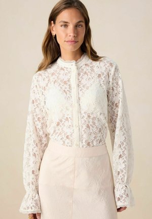 Witte kanten blouse met lange mouwen, hoge hals en knoopsluiting. Bevat bloemige patronen en geribde manchetten. Gepaard met een lichtroze rok.
