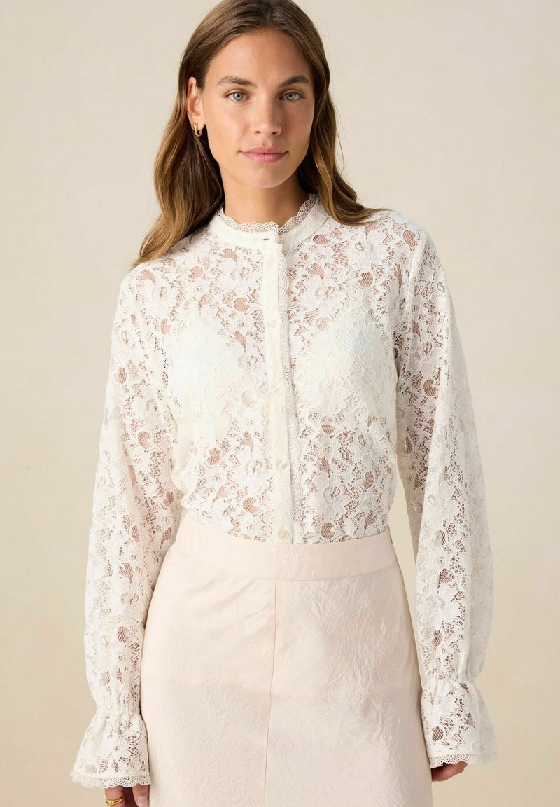 White lace blouse with long sleeves, high neck, and button closure. Features floral patterns and frilled cuffs. Paired with a light pink skirt.