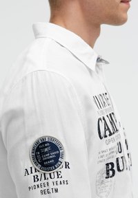 Weißes Baumwollhemd mit Textdruck und einem runden navyblauen Aufnäher auf dem linken Ärmel. Klassischer Kragen und strukturierter Stoff.