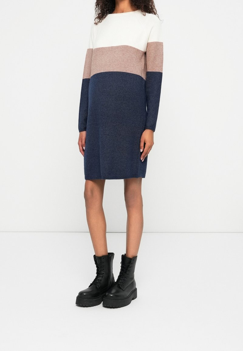 Femme portant une robe en maille à manches longues color block blanche, beige et bleu marine, associée à des bottines à plateforme noires lacées, debout contre un mur blanc.
