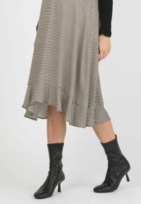 Schwarz-creamfarbenes, gemustertes Kleid mit asymmetrischem Saum. Das Model trägt enganliegende schwarze Stiefeletten. Strukturierter Stoff und Rüschendetail am Saum.