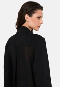 Blazer nero con design strutturato, caratterizzato da una parte posteriore parzialmente trasparente, colletto classico e dettaglio con bottoni sulle maniche.