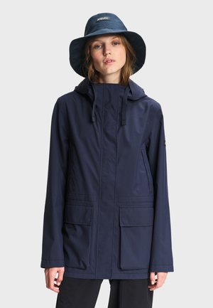 Frau trägt eine marineblaue Regenjacke mit Vorderentaschen und einen passenden breitkrempigen Hut vor einem einfarbigen hellen Hintergrund.