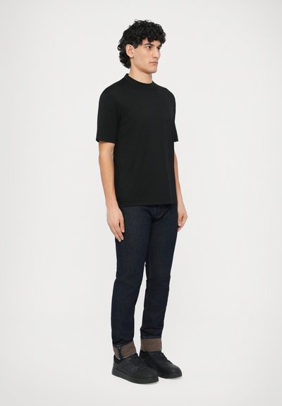 Fekete rövid ujjú póló kerek nyakkal, sötétkék farmernadrággal, fekete tornacipővel és visszahajtott szegéllyel, amely barna bélést mutat.